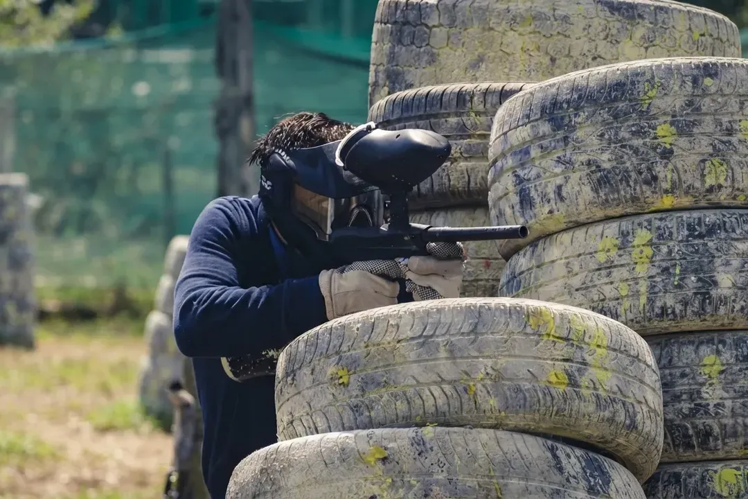 Paintball player in a mask aiming a marker from behind a stack of paint-splattered tyres